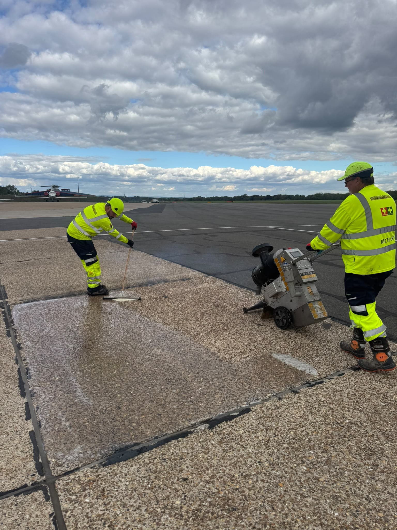 Two Shepherd and Sons operatives in hi-vis preparing a concrete bay on an airfield apron, one working a surface planer and one spreading material with a squeegee, with an aircraft parked in the background